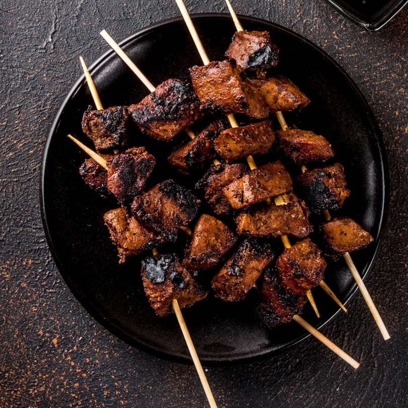Top-down view of several Alice's Bar-B-Que skewers with charred meat pieces on a black plate.