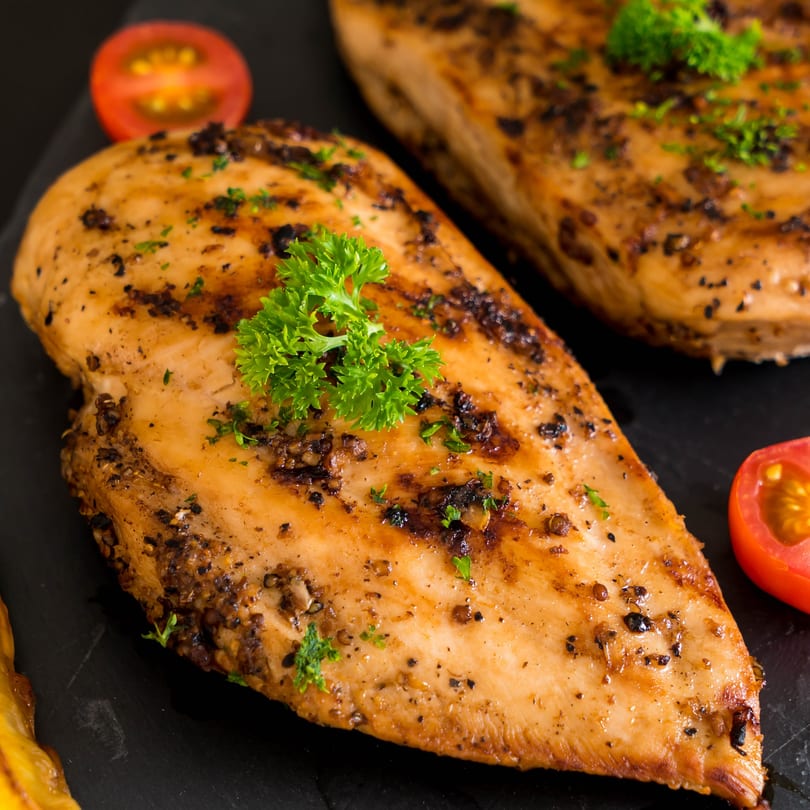 Two grilled chicken breasts seasoned with herbs and spices, garnished with parsley and tomato slices.